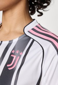 Bijela sportska majica s crnim i ružičastim trakama, koja ima izvezeni logo s tri zvjezdice i kontrastni rub oko ovratnika.