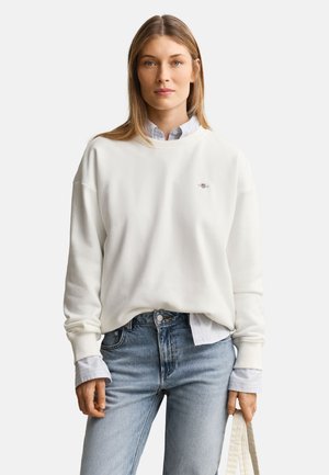 Blond kvinde iført hvid sweatshirt over en knaplukket skjorte, lyseblå jeans, der holder en cremefarvet shopper mod en ensfarvet baggrund.