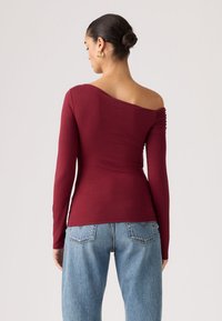 Μπορντό μπλούζα με μακριά μανίκια, ασύμμετρο ντεκολτέ και ρεβάνς σε έναν ώμο, σε συνδυασμό με ανοιχτόχρωμο τζιν παντελόνι.