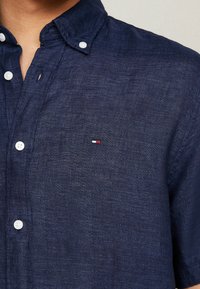 Navy blue button-up shirt in a textured fabric, featuring a collar, white buttons, and a small red, white, and blue logo on the chest.