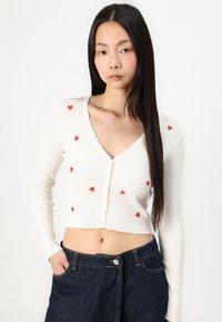 Cardigan bianco a coste con taglio corto, maniche lunghe e piccoli dettagli a forma di cuore rosso. Chiusura frontale con bottoni che aggiunge particolare.