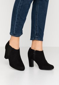 Černé semišové kotníkové boty s špičatou špičkou a 7 cm podpatkem. Postranní elastické pásky a zadní zip zvýrazňují design. Nosené s tmavými džínami.