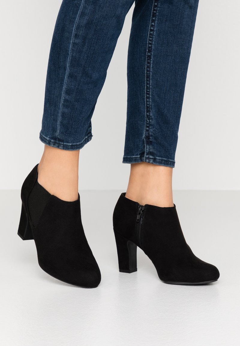Černé semišové kotníkové boty s špičatou špičkou a 7 cm podpatkem. Postranní elastické pásky a zadní zip zvýrazňují design. Nosené s tmavými džínami.