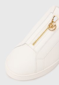 White sneaker with gold zipper and Michael Kors logo pull on top, showing detailed stitching and smooth material.