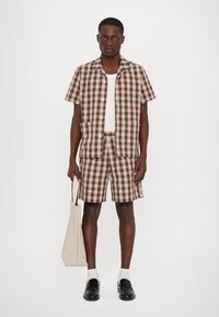 Checkered short-sleeve shirt and shorts set in brown and white, featuring a button front, two pockets, white undershirt, and black shoes.