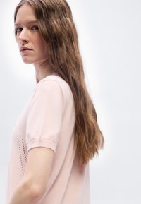 Νεαρή γυναίκα με μακριά καστανά μαλλιά και ανοιχτό ροζ μπλουζάκι με κοντά μανίκια κοιτάει πλάγια, πάνω σε λευκό φόντο.