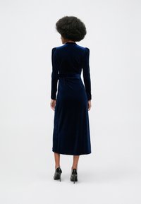 Ženska z afro frizuro nosi midi obleko z dolgimi rokavi v mornarsko modri barvi iz žameta in črne visoke pete, stoji s hrbtom proti kameri.