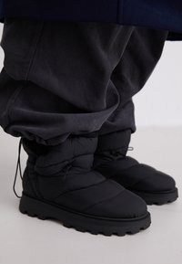 Black padded winter boots with a rubber sole, textured stitching, and drawstring closure. Paired with loose, dark grey corduroy pants.
