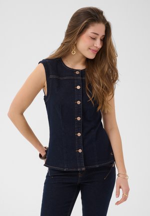 Kvinde med langt hår iført mørk denim top uden ærmer med knapper og matchende mørke denim bukser, poserer med lukkede øjne og let smil.