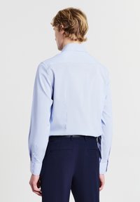 Uomo dai capelli biondi che indossa una camicia a maniche lunghe azzurro chiaro e pantaloni blu scuro, in piedi di spalle su uno sfondo bianco.