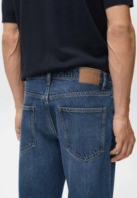 Jeans negri închis, din denim, având o etichetă din piele maro pe spate, cinci buzunare și detalii de cusătură vizibile.