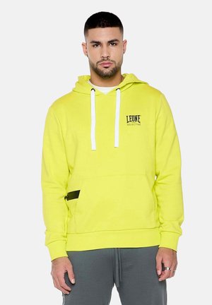 Man met kort haar en baard, die een helder gele hoodie met witte trekkoordjes en grijze broek draagt tegen een witte achtergrond.