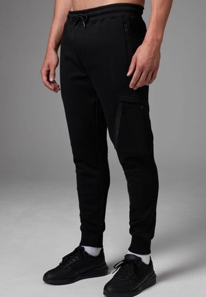 Svarta joggingbyxor i mjukt material, med sidofickor med dragkedja, en figurnära design och ribbade muddar. Bärs med svarta träningsskor.