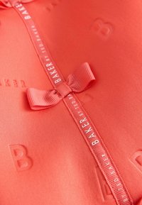 Raudonos spalvos audinys su iškiliais užrašais ir juostele viduryje, ant kurios yra balta etiketė su užrašu "BAKER by TED BAKER".