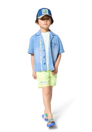 Denim short-sleeve shirt, light blue, with colorful text. Light green shorts with a wave design. Blue sandals with logo accents.