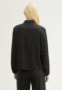 Blusa negra de manga larga con bolitas blancas, tejido suave, detalle en el cuello y un corte relajado. Costura trasera visible y puños abotonados.