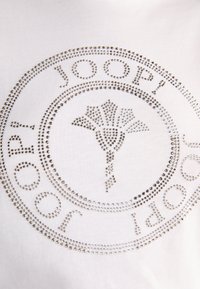 Camiseta blanca con brillantes plateados en un patrón circular, que muestra el nombre de la marca "JOOP!" y un diseño estilizado de una flor en el centro.