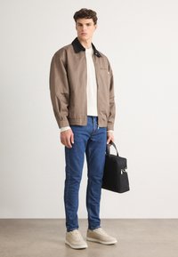 Brązowa lekka kurtka z czarnym kołnierzem, biała dziana sweter, niebieskie wąskie dżinsy i beżowe sneakersy. Trzyma czarną torbę. Prosty design.