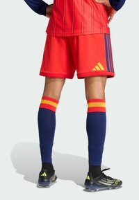 Punaiset urheilushortsit yhdistettynä sinisiin sukkiin, joissa on keltaisia ja punaisia raitoja. Mustat ja keltaiset nappikset, joissa on teksturoitu pinta, nähtynä takaa.