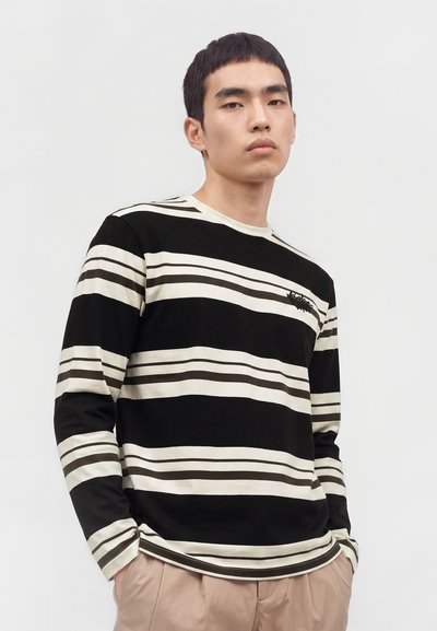 Jeune homme portant une chemise à manches longues rayée noire et blanche avec un pantalon beige, debout devant un fond clair uni.