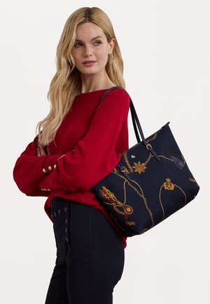 Femme blonde portant un pull rouge porte un grand sac cabas noir avec des motifs de chaînes en or et d'emblèmes sur l'épaule.
