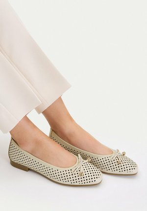Beżowe baleriny z perforacjami i małymi kokardkami, noszone na boso z jasnymi, krótkimi spodniami.