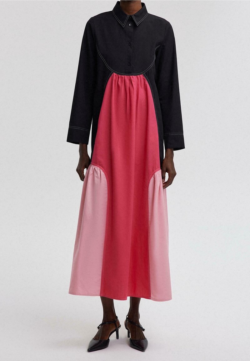 Femme portant une longue robe color block avec un haut et un col noirs, un panneau central rouge et des panneaux latéraux rose clair, associée à des talons noirs.