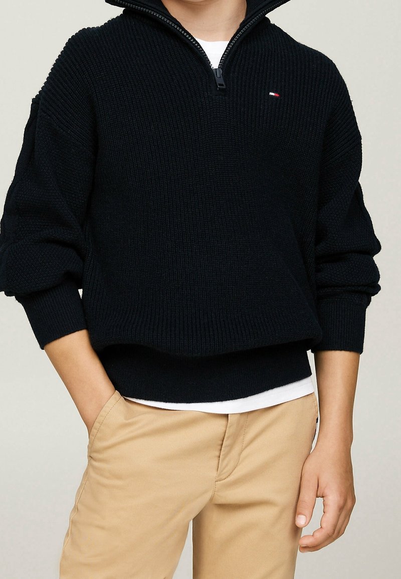 Enfant portant un pull en maille côtelée noir avec une fermeture éclair au quart et un pantalon beige, debout, une main dans la poche, sur un fond uni.