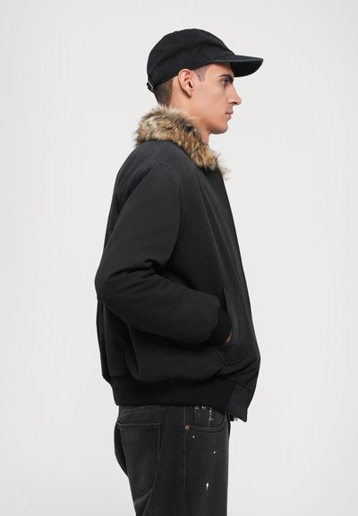 Svart bomberjacka med pälskrage, med avslappnad passform och sidofickor. Bärs med en svart keps och mörka jeans, profilvy.