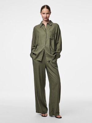 Vrouw staat met handen in de zakken, draagt een losse olijfgroene blouse met knopen en wijde broekspijpen, gecombineerd met zwarte sandalen met open tenen.