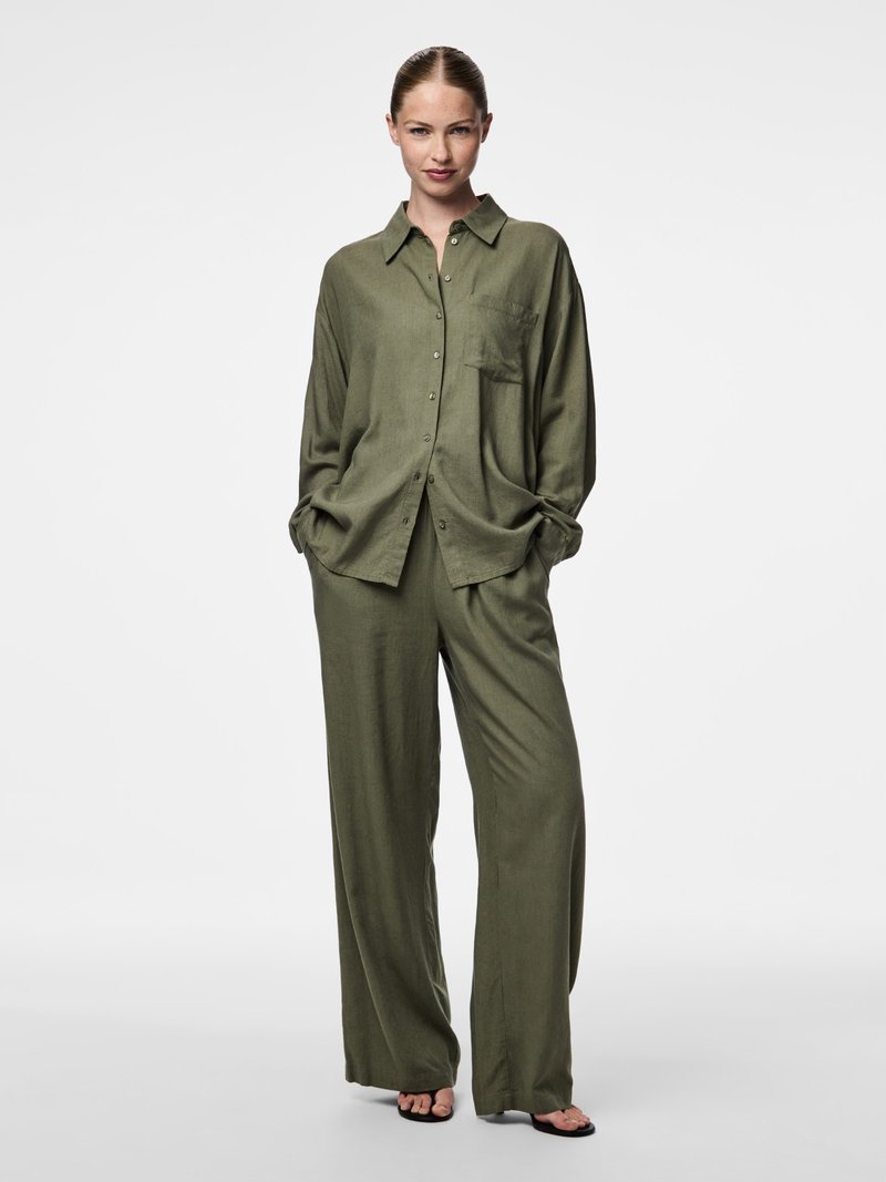 Kvinne står med hendene i lommene iført løs olivengrønn knappeskjorte og vidbenede bukser, kombinert med svarte sandaler med åpne tær.