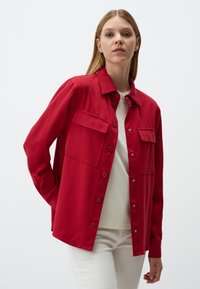 Chemise rouge à boutons en tissu doux, dotée de deux poches poitrine, de détails en métal argenté et d'une coupe décontractée. Portée par-dessus un haut blanc.