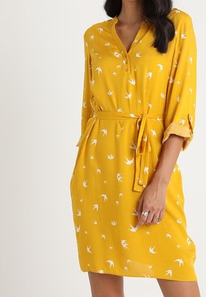 Żółta sukienka do kolan z białymi nadrukami ptaków, rękawami do łokcia, rozpinanym dekoltem i przewiązanym paskiem w talii.