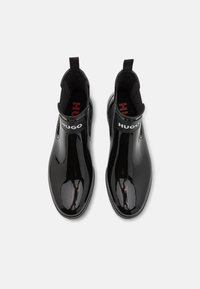 Par de botas negras brillantes hasta el tobillo con lados elásticos, tiradores y etiqueta blanca de la marca "HUGO" en la parte superior de cada bota.