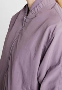 Kvinna iklädd en mauvafärgad zip-up-jacka med ribbat krage, visad från halsen till överkroppen mot en neutral bakgrund.