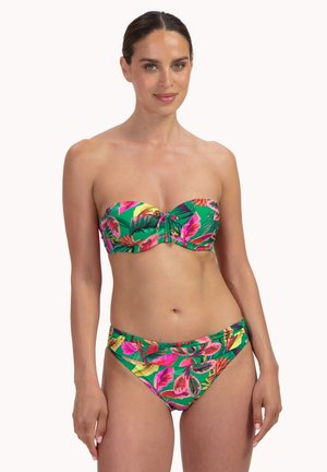 Mujer con bikini sin tirantes de estampado tropical con patrones florales en verde, rosa y naranja, sonriendo y de pie frente a un fondo liso.