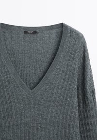 Pull en maille gris avec col en V et motif torsadé, comportant une petite étiquette noire au niveau du col.