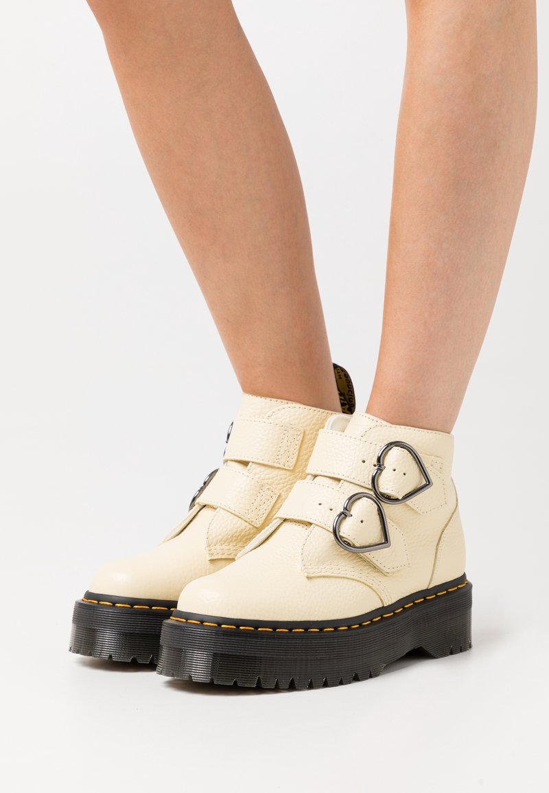 Bottines crème avec semelles épaisses noires et deux boucles en métal en forme de cœur sur chaque bride, portées sur jambes nues.