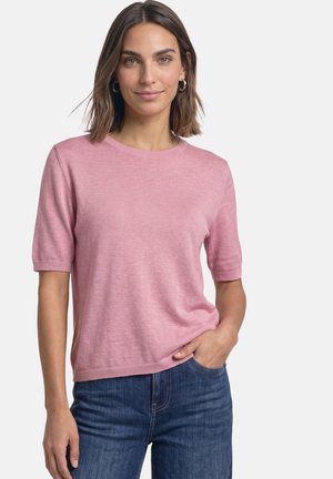 Femme aux cheveux bruns mi-longs portant un pull rose à manches courtes et un jean bleu, souriant avec une main dans la poche.