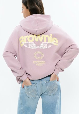 Mujer vistiendo una sudadera con capucha rosa con la palabra "Brownie" y gráficos de alas, combinada con vaqueros azul claro que tienen diseños de alas tachonadas en los bolsillos traseros.