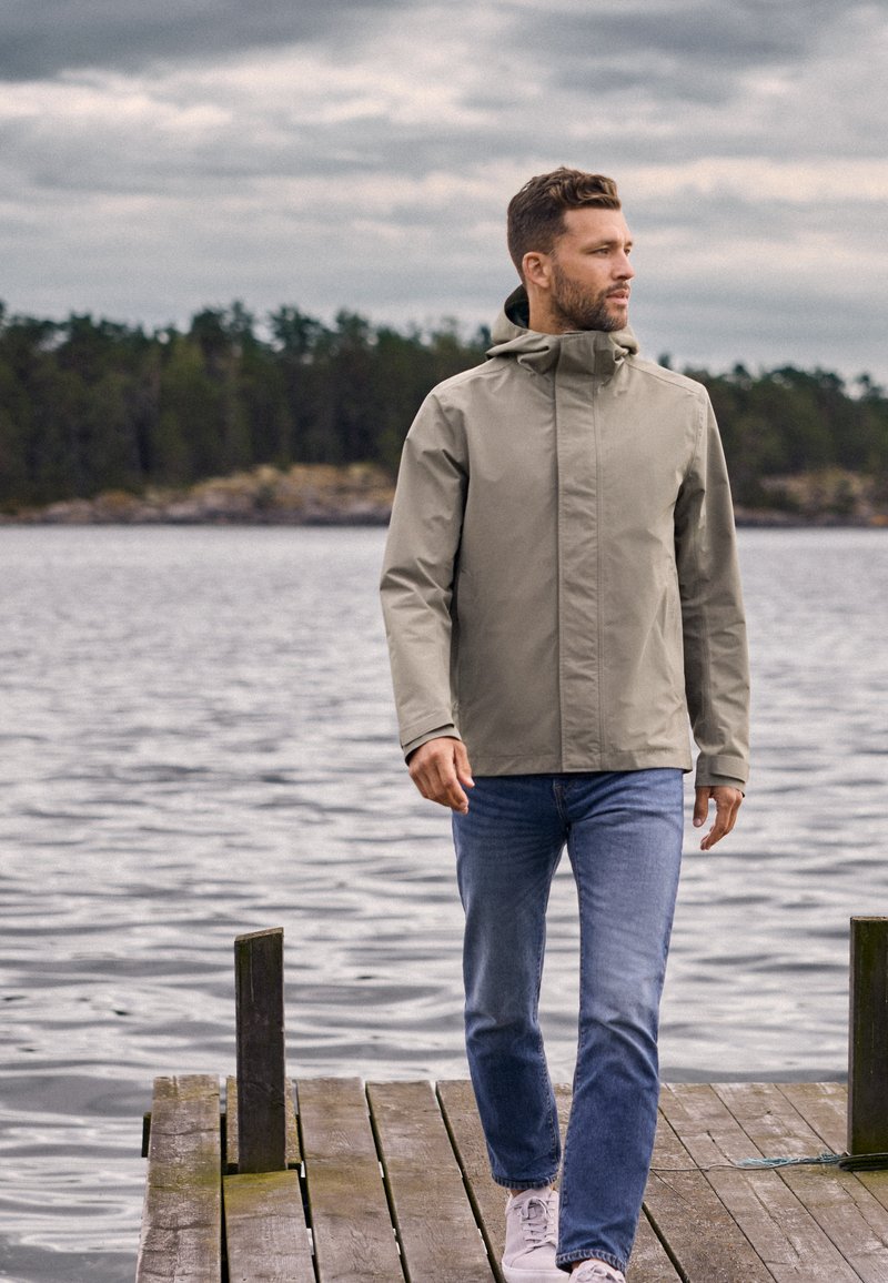 Mann trägt beige Jacke und blaue Jeans, geht auf einem hölzernen Steg an einem ruhigen See, im Hintergrund Wald und bewölkter Himmel.