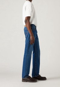 Jeans rectos azules con cintura alta, tela de mezclilla texturizada y una etiqueta de cuero en la parte trasera. Combinados con una camiseta blanca básica y zapatos oscuros.