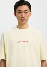 Mladić s ravnom crnom kosom, nosi krem bijelu majicu s crvenim natpisom "HÔTEL AMOUR" na jednobojnoj bijeloj pozadini.