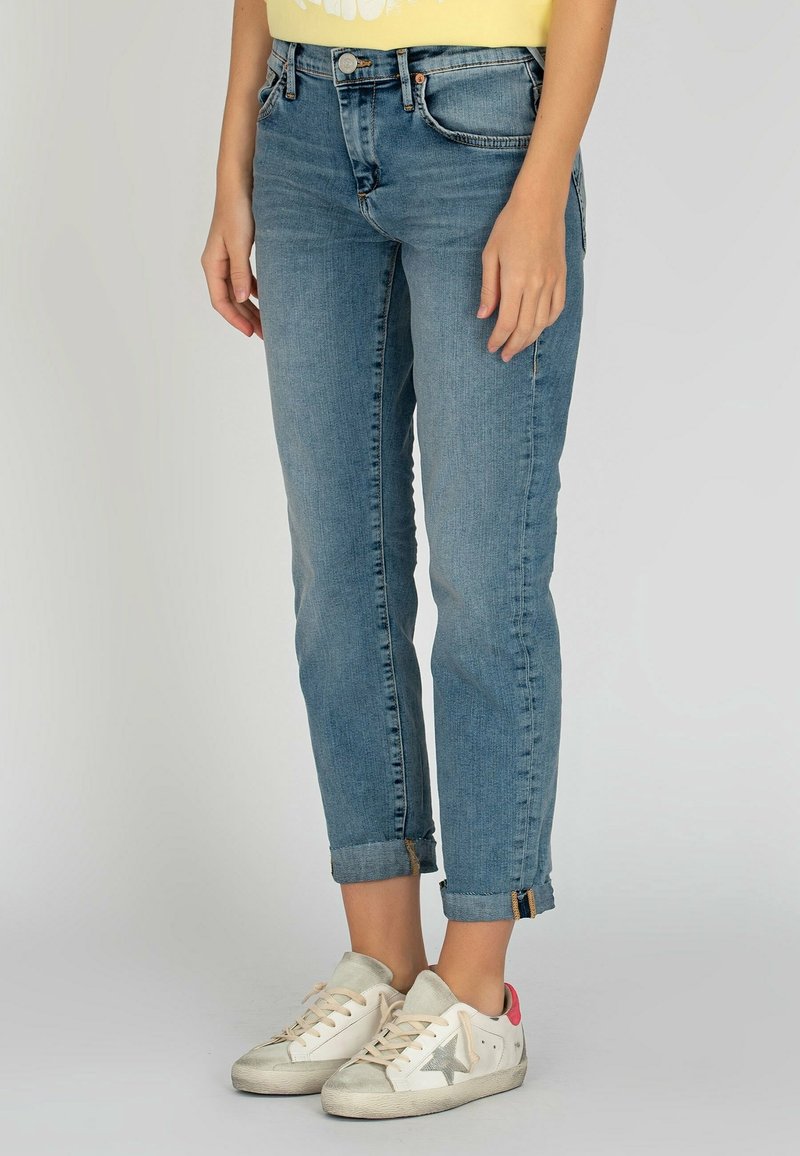Personne portant un jean bleu clair retroussé et des baskets blanches avec des accents étoilés gris, debout devant un fond neutre.