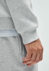 Halli sweatshirt kangas ribbed mansettidega, näidates pehmet tekstuuri. Käsi hoiab riietust, paljastades sujuva naha tooni.