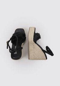 Schwarze Wedgesandalen mit einem geflochtenen Riemendesign. Aus Leder gefertigt und mit einer gewebten Jutebasis ausgestattet. Glatte Textur mit einem leichten Schimmer.