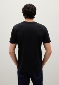 Uomo con capelli ricci scuri che indossa una maglietta nera a maniche corte semplice e jeans scuri, in piedi con le mani in tasca di fronte a una parete grigia chiaro.