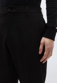 Čierne na mieru šité nohavice z hladkej tkaniny, s plochým predným dielom, skrytým zapínaním na gombíky a bočnými vreckami. Na obrázku je ruka položená na tkanine.