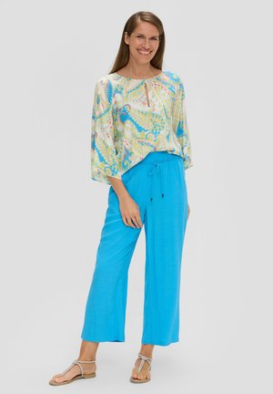 Bluse mit buntem Paisley-Muster in Gelb, Blau und Grün; lockere Passform. Helle blaue, weit geschnittene Hose mit Kordelzugbund. Sandalen.