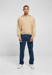 Felpa beige con cappuccio, tasca frontale e chiusura a mezza zip, abbinata a jeans blu scuro e sneakers bianche. Design semplice e casual.